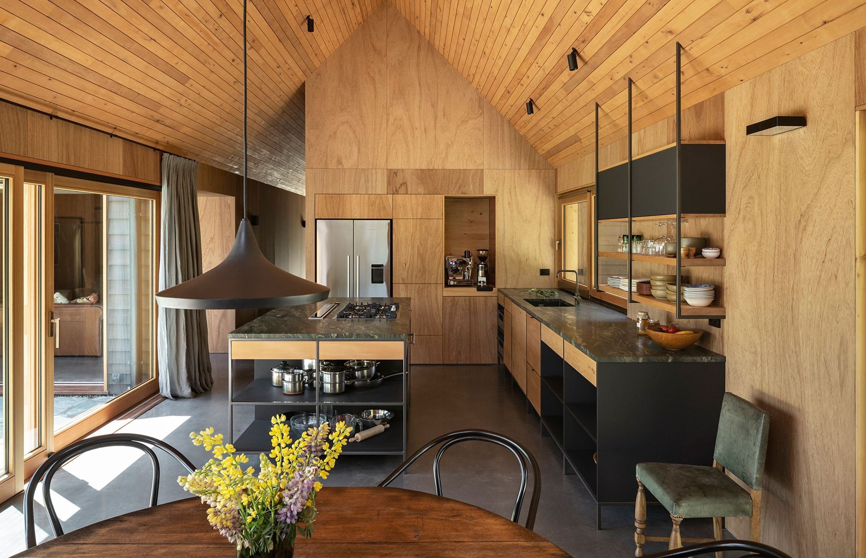 The open-plan kitchen has a wall of plywood cabinetry and an island and bench constructed in mild steel and recycled rimu, with a striking verde marble benchtop, like pounamu.