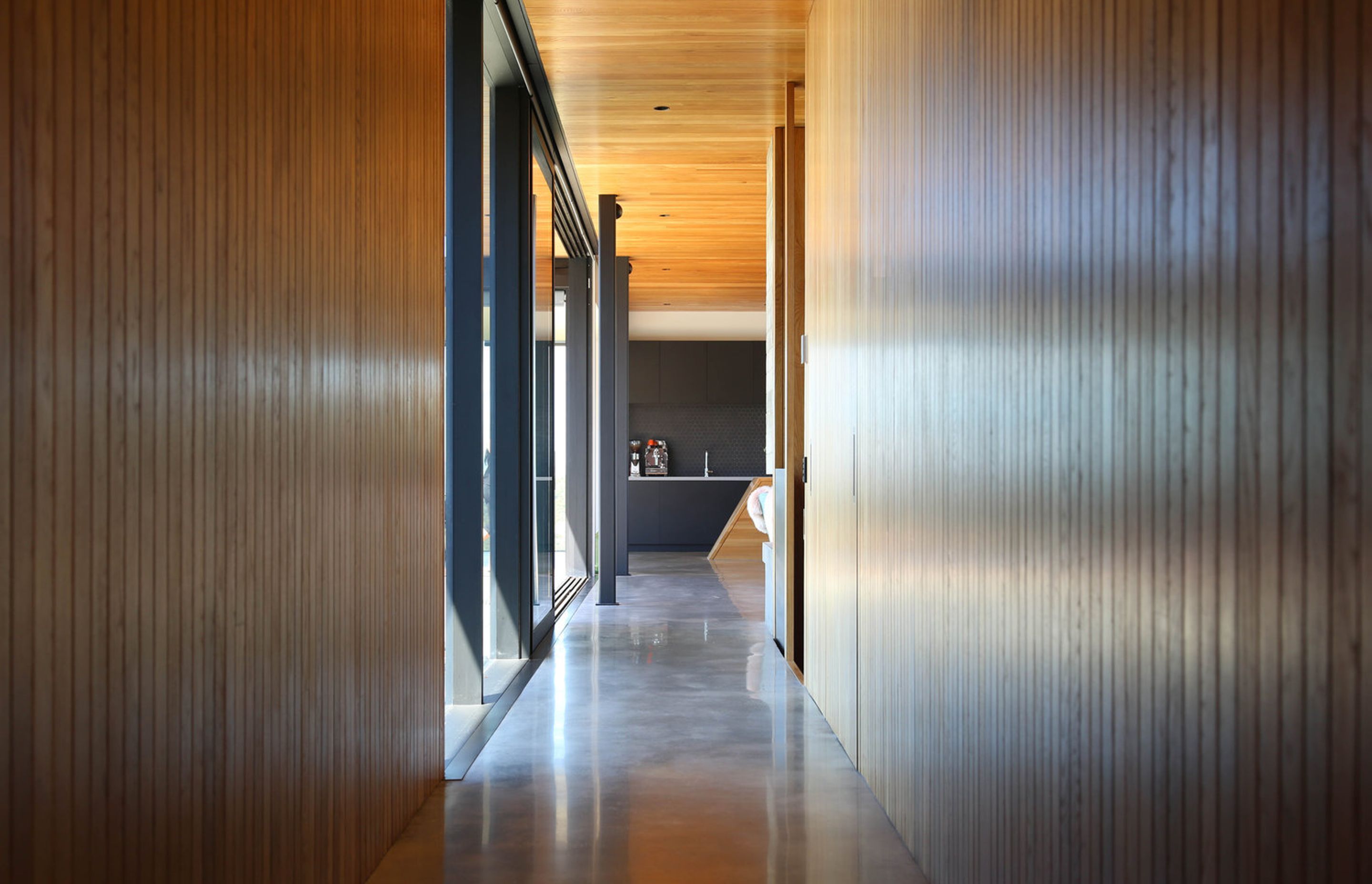The natural oak-lined corridor from the entry to the kitchen has a polished concrete floor and black aluminium joinery.