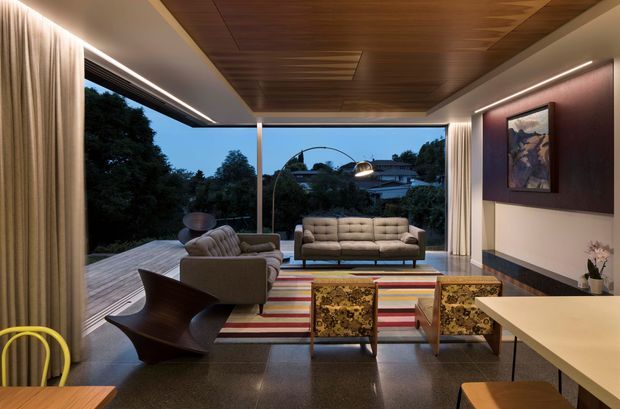 A refined contemporary living room opening onto a timber deck, with full-height glazing and curtains framing a leafy suburban outlook. The room features a warm timber-lined ceiling, dark polished flooring, a large striped rug, a neutral sofa and armchairs, patterned accent chairs, sculptural lounge seating, integrated linear lighting, and a dark feature wall with artwork.