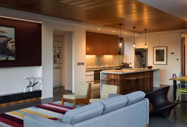 Open-plan contemporary living, dining and kitchen interior with a warm timber-lined ceiling, white walls, dark flooring, and strong mid-century-inspired furnishings. A large grey sectional sofa faces a dark feature wall and fireplace area. The kitchen includes a timber island with integrated seating, dark benchtops, and a colourful patterned splashback, while pendant lights hang above the island. A dining area with colourful chairs is visible to the right, and built-in shelving is recessed near the kitchen.