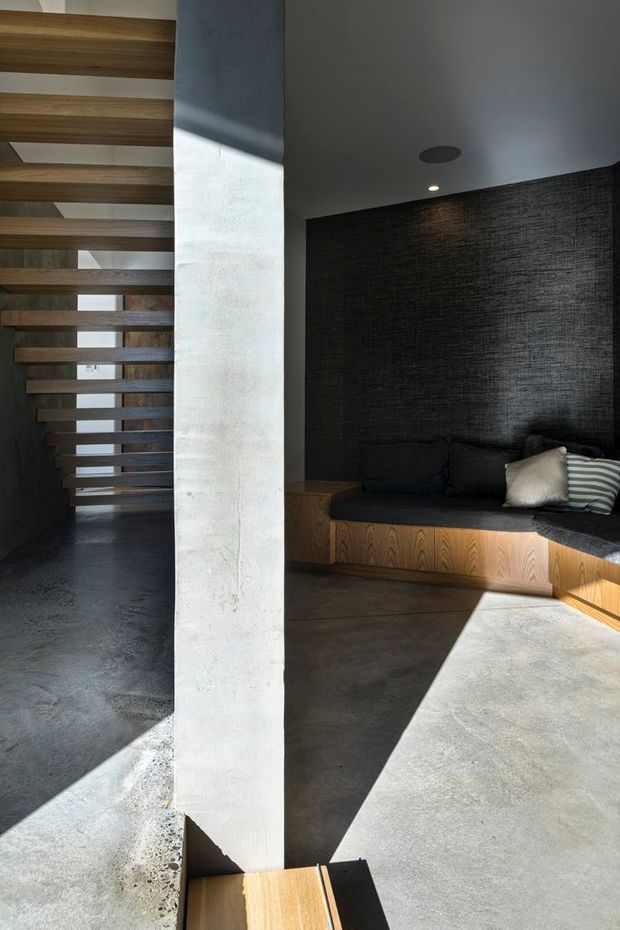 A dramatic modern interior featuring a floating timber stair with open risers on the left, a substantial pale concrete structural column in the centre, and a recessed lounge nook on the right. The lounge has a built-in timber bench with dark cushions against a textured charcoal wall. The floor and ceiling are dark and concrete-toned, with strong sunlight creating angular shadows across the space.