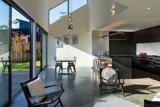 A bright, open-plan contemporary interior combining a dining area, kitchen and living space. Full-height glazed sliding doors open to a timber deck and garden, while a double-height ceiling brings in abundant daylight. The kitchen features dark built-in cabinetry, a long island with stools, integrated appliances and pendant lighting. Polished concrete flooring, neutral walls, framed artwork and mid-century-inspired lounge chairs complete the space.
