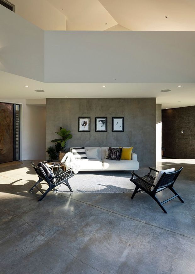A spacious contemporary living room with a polished concrete floor, exposed concrete feature wall, white sofa, black leather-and-cane lounge chairs, and framed artwork. The room has a high ceiling with a partial upper-level void, recessed lighting, abundant natural light, and dark textured wall surfaces in an adjacent area.