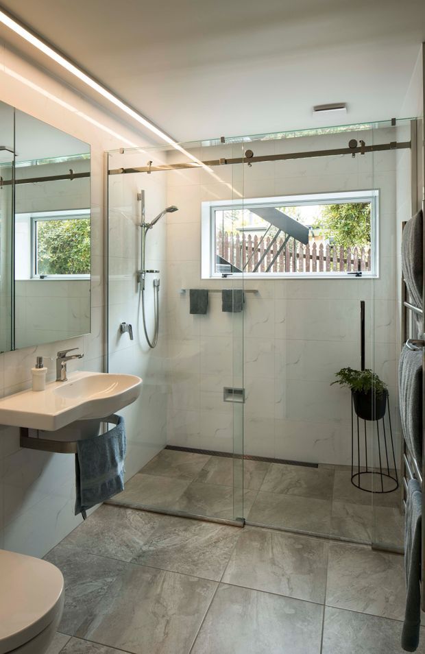 A bright contemporary bathroom featuring a large frameless glass walk-in shower with dark exposed sliding-door hardware, wall-mounted shower fittings, pale marble-look wall tiles, and large grey stone-look floor tiles. A white wall-mounted basin with chrome tapware sits beside a full-height mirror, with folded towels on a vertical rail and a potted plant adding greenery. Natural light enters through a horizontal window overlooking a fenced garden.