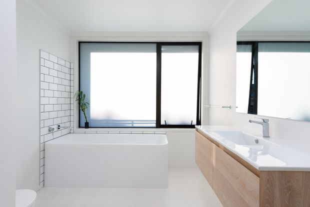 Bright contemporary bathroom featuring a freestanding white bathtub beneath large black-framed frosted windows, white rectangular wall tiles with dark grout, and a light timber vanity with a white benchtop and integrated basin. A large mirror reflects the window and vanity area.