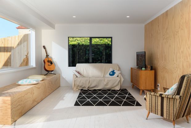 Bright, compact living room with white walls and pale timber flooring, featuring a plywood-clad built-in window bench, plywood feature wall, cream slipcovered sofa, vintage wooden sideboard, patterned rug, and upholstered armchair. A guitar and television add residential character, while large windows provide natural light and views toward greenery.