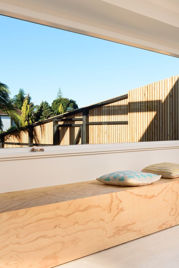 A bright, minimalist interior looking through a large opening toward a timber-clad outdoor structure. The foreground features a built-in plywood bench or platform with a pale wood grain finish, white walls and trim, and cushions. Strong sunlight and the visible timber fence-like facade create a warm, contemporary architectural setting.