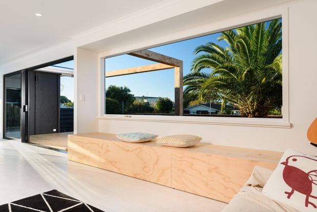 Bright contemporary interior featuring a large picture window overlooking lush palm trees and neighbouring rooftops. A built-in light timber window bench or cabinet runs beneath the glazing, paired with a cream upholstered sofa and patterned cushions. Black-framed glazed doors open to the exterior, while a black-and-white geometric rug adds contrast.