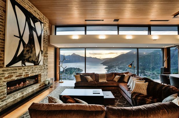 A warm, sophisticated living room with a large brown sectional sofa arranged around a low rectangular coffee table and a linear fireplace set into a rough stone feature wall. The room has a timber-lined ceiling, exposed brick and stone surfaces, expansive black-framed glazing, and dramatic mountain-and-water views beyond. A mezzanine-like upper opening and clerestory glazing contribute to the spacious architectural composition.