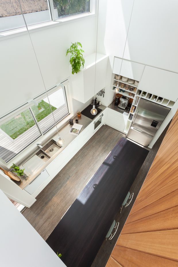 Bright contemporary kitchen viewed from above, featuring white cabinetry, a large dark timber island, stainless-steel appliances, a double sink beneath expansive windows, pendant lighting, and warm timber wall panelling. The kitchen opens visually toward an outdoor deck and lawn, with a tall leafy indoor plant adding greenery.