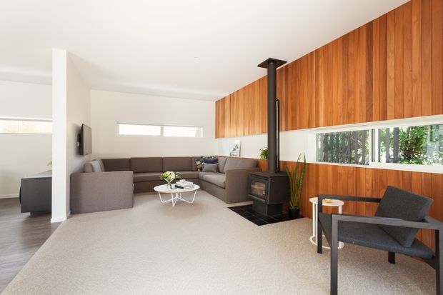 Bright, open-plan living room with a large brown sectional sofa, pale carpet, white walls, a freestanding black wood-burning stove, and a dramatic full-height timber feature wall incorporating a long horizontal window. A dark timber-framed lounge chair and small side table sit in the foreground, with a glimpse of the adjacent kitchen or dining area at left.