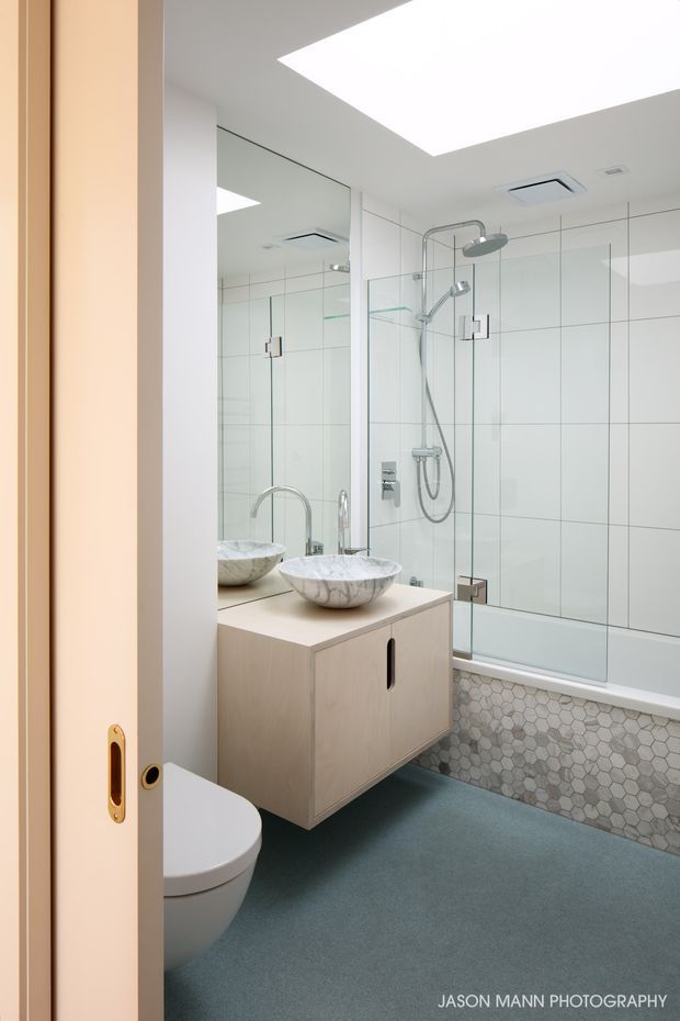 Bright, compact contemporary bathroom viewed through a partially open sliding door. The room features a floating light-wood vanity with a pale stone or solid-surface benchtop, vessel basin, large wall mirror, frameless glass shower screen, chrome shower fittings, white wall tiles, a bathtub with a patterned hexagonal-tile front, and a blue-grey floor. A skylight or large ceiling light opening provides strong overhead illumination.