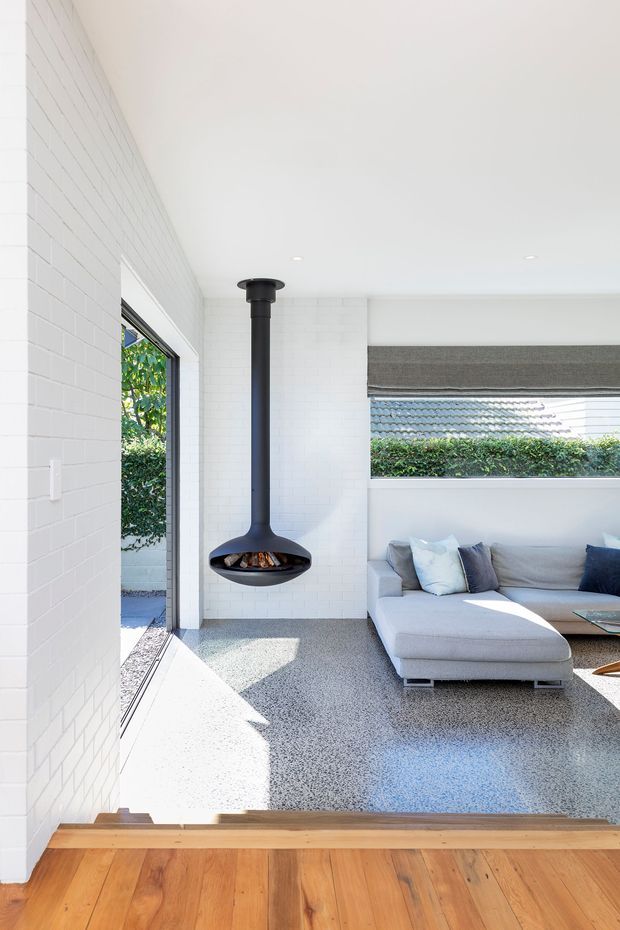 Bright contemporary living room with white painted brick walls, polished terrazzo flooring, a suspended black fireplace, and a light grey sectional sofa. Timber flooring and steps lead into the room, with large glazing, a roller blind, and greenery visible outside.