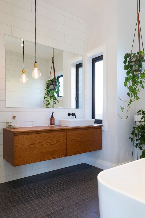Bright contemporary bathroom with a wall-mounted natural timber vanity, white vessel basin, matte black tapware, large frameless mirror, white rectangular wall tiles, dark small-format floor tiles, black-framed windows, pendant lights, hanging plants, and a partially visible freestanding bathtub.