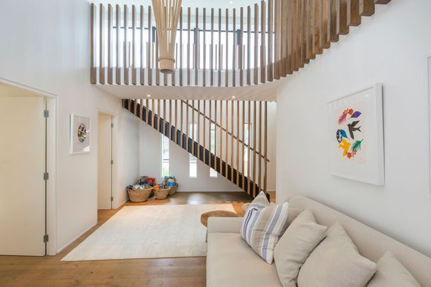 Bright contemporary interior showing a living room beneath a sculptural timber staircase and curved upper balustrade. White walls, pale upholstered sofa, warm timber flooring, and extensive vertical timber slats create an airy, refined space. Artwork and decorative baskets add colour and texture.