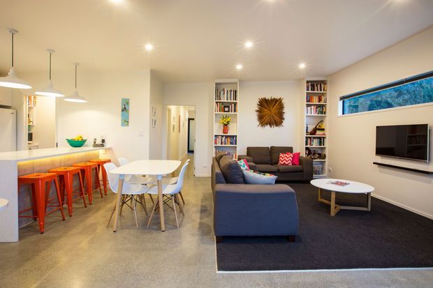 Open-plan contemporary living area combining a kitchen with breakfast bar, dining zone, and lounge. The space features polished concrete flooring, white walls and ceiling, integrated shelving filled with books, a dark sectional sofa, colourful bar stools, pendant lighting, recessed downlights, and a narrow horizontal clerestory window above the wall-mounted television.