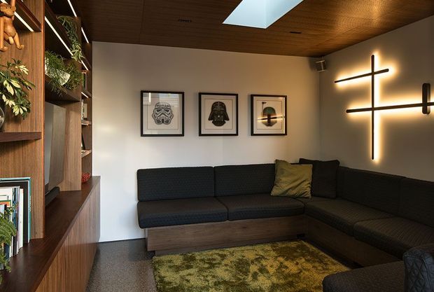 A compact, moody living room with a dark L-shaped sectional sofa, timber built-in shelving and cabinetry, framed graphic artwork, and a glowing linear wall light. The room has a richly stained timber ceiling with a skylight, dark textured flooring, and a patterned area rug, creating a warm contemporary media-lounge atmosphere.