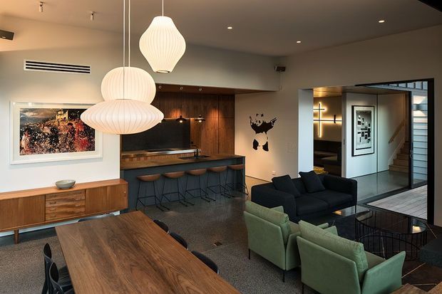 A warm, mid-century-inspired open-plan living and dining area with a built-in timber bar and kitchen joinery. The foreground features a large timber dining table, while the lounge has black and muted green upholstered armchairs around a low coffee table. Three sculptural white pendant lights hang above the dining and bar areas. Dark timber wall panelling, integrated shelving, framed artwork, and a glazed opening toward a hallway and stairway create a refined retro interior.
