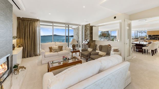 Bright, elegant open-plan living room with cream upholstered sofas, striped armchairs, a timber coffee table, and a textured stone fireplace wall. Large windows and glazing provide a waterfront view, while the adjoining dining and kitchen area is visible to the right. The interior has a refined coastal feel with neutral finishes, layered curtains, and abundant natural light.