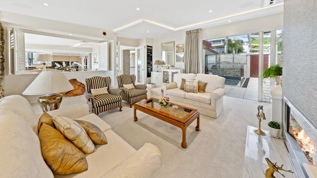 Bright, spacious formal living room with cream upholstered sofas, striped armchairs, a timber and glass coffee table, and a marble-look fireplace surround. White built-in cabinetry, decorative wall mirrors, framed artwork, recessed lighting, and large glazed doors create an elegant, refined interior with views to an outdoor courtyard.