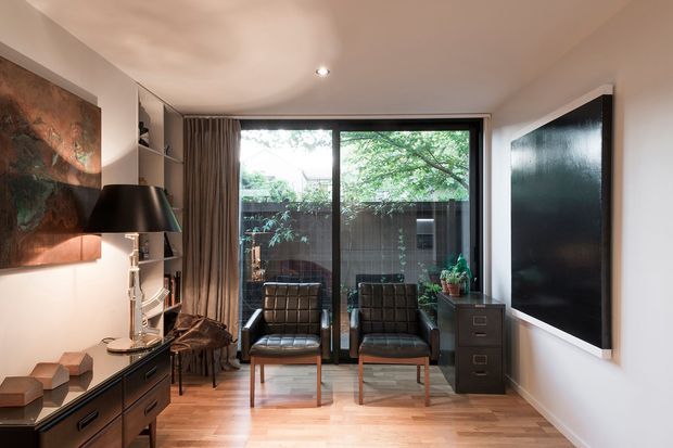 A compact, refined living room or sitting area with warm timber flooring, white walls, a large black-framed glazed door or window opening to a planted courtyard, two dark leather lounge chairs, and a dark timber sideboard. Built-in shelving, full-height curtains, a large black wall-mounted panel, artwork, and a sculptural table lamp create a sophisticated contemporary interior.