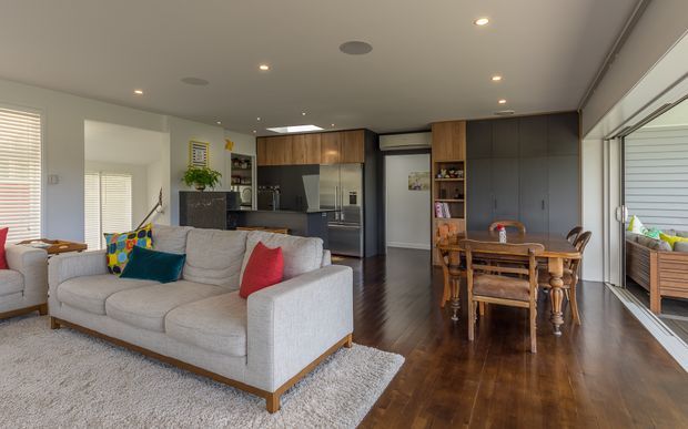 Open-plan contemporary living and dining area with a neutral sectional sofa, dark timber flooring, integrated kitchen cabinetry, and a compact timber dining table. Large glazed doors or windows open to the exterior, while recessed ceiling lights and dark built-in joinery create a clean, modern interior.