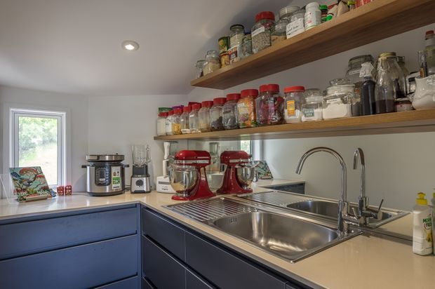 Compact, well-organised kitchen with deep blue-grey flat-panel base cabinetry, pale benchtops, a large stainless-steel double sink, and open timber shelves displaying jars and pantry goods. Red stand mixers and countertop appliances sit beneath recessed ceiling lighting, with a window providing natural light.