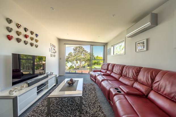 Bright contemporary living room with a large red-brown leather sectional sofa, white built-in media cabinetry, wall-mounted television, central coffee table, and textured dark rug. Full-height sliding glass doors open to an outdoor area, while decorative wall objects and framed artwork add visual interest. A wall-mounted split-system air conditioner is visible above the sofa.