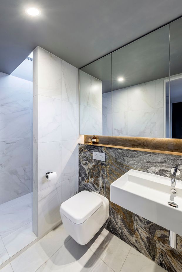A compact, luxurious contemporary bathroom featuring large white marble-look wall and floor tiles, a wall-hung toilet, a white rectangular vessel basin, a dramatic dark veined stone vanity wall and benchtop, full-height mirrored cabinetry, and recessed ceiling lighting. A glass shower enclosure is visible to the left.