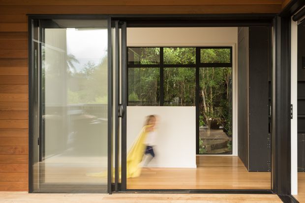 View through a large open glazed sliding doorway into a warm timber-lined interior. The space features honey-toned timber flooring and wall cladding, dark aluminium-framed glazing, and a leafy garden visible beyond the rear windows. A blurred person is moving through the room, with minimal furnishings visible.