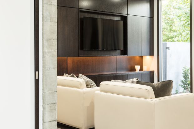 A contemporary living room viewed from an entry or adjacent space, centred on a large dark timber built-in media wall with an inset television, illuminated display niche, and low console. Cream sofas with patterned cushions face the media unit. A raw concrete structural column and tall glazing or an open doorway at the side add contrast, while concealed lighting creates a refined, warm atmosphere.