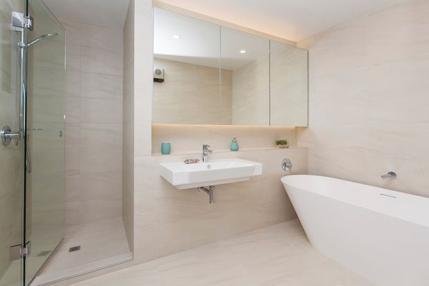 A bright, contemporary bathroom featuring a frameless glass walk-in shower, wall-mounted white basin, mirrored wall cabinetry, a freestanding bathtub, and pale beige stone-look wall and floor tiles. Recessed lighting and a softly illuminated vanity shelf create a clean, serene atmosphere.