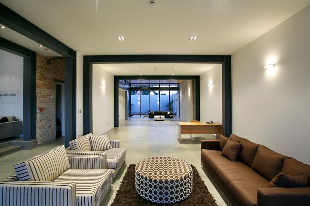 A spacious contemporary lounge and circulation area with a long axial view toward a glazed courtyard or garden. The room features polished concrete flooring, white walls, dark charcoal structural openings and trims, exposed brickwork at the left, striped upholstered lounge chairs, a brown sofa, a patterned round ottoman, and a simple timber console. Recessed ceiling lights and wall sconces provide clean modern illumination.