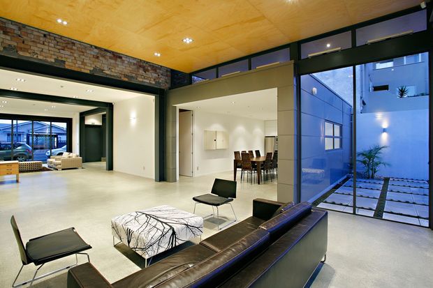 Open-plan contemporary living area with a lounge seating arrangement in the foreground, dining area beyond, and extensive full-height glazing opening views to a minimalist courtyard. The space features a polished light floor, dark steel-framed glazing and joinery, exposed brickwork, and a warm plywood ceiling with recessed lighting.