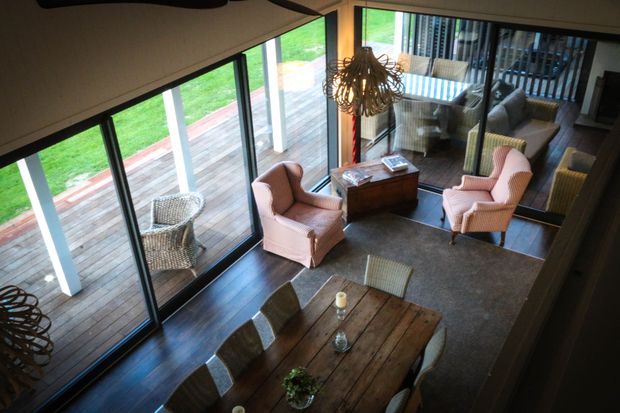 A warm, double-height living and dining area viewed from above, with extensive floor-to-ceiling glazing opening onto a timber deck and lawn. The room contains a rustic timber dining table with upholstered chairs, two pink armchairs, a small wooden side table, dark timber flooring, and a sculptural pendant light. Reflections in the glazing reveal additional seating and the deck beyond.