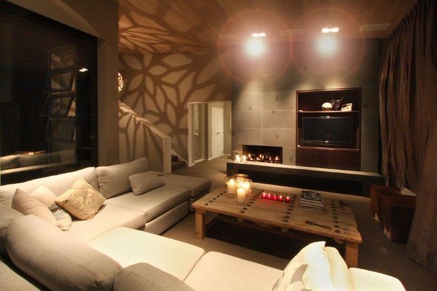 A moody, warmly lit living room with a large cream sectional sofa arranged around a rustic timber coffee table. The room features a dark tiled feature wall with a built-in fireplace and recessed media cabinetry, patterned organic wall and ceiling shadows, dark curtains, and a large window or glazed opening on the left. Candles and a fire add a cozy atmosphere.