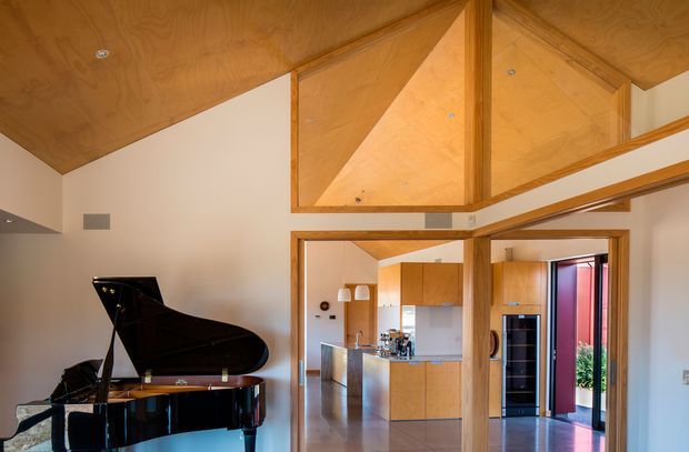 Interior view of an open-plan contemporary home featuring a grand piano in the foreground, extensive warm timber-lined sloped and vaulted ceilings, and a kitchen visible through a wide opening. The kitchen has timber cabinetry, a central island, stainless-steel appliances, and a polished floor. Glazed doors at the rear open toward an outdoor area.