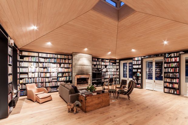 A warm, expansive home library and reading room with floor-to-ceiling dark shelving filled with books, timber flooring, and a striking timber-lined faceted ceiling with a small skylight. A central stone or concrete fireplace is set between the bookshelves, with sofas, leather lounge chairs, a timber coffee table, and reading seating arranged around it. Glazed French doors and additional built-in shelving line the rear wall.