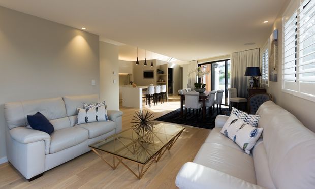 Bright open-plan living and dining area with a connected kitchen in the background. Two white sofas face a glass coffee table with a geometric metal base, while a dark rug anchors the dining table and upholstered chairs. Neutral walls, pale timber flooring, recessed ceiling lighting, pendant lights, full-height curtains, and white plantation shutters create a refined, comfortable interior.