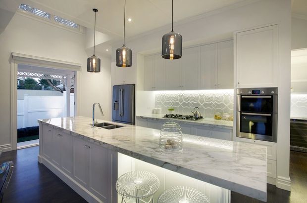 Bright, refined white kitchen with a large marble-look island, integrated white cabinetry, stainless-steel appliances, patterned tile splashback, pendant lighting, and dark timber flooring. The island includes a sink and seating, while a glazed door opens to an outdoor area.