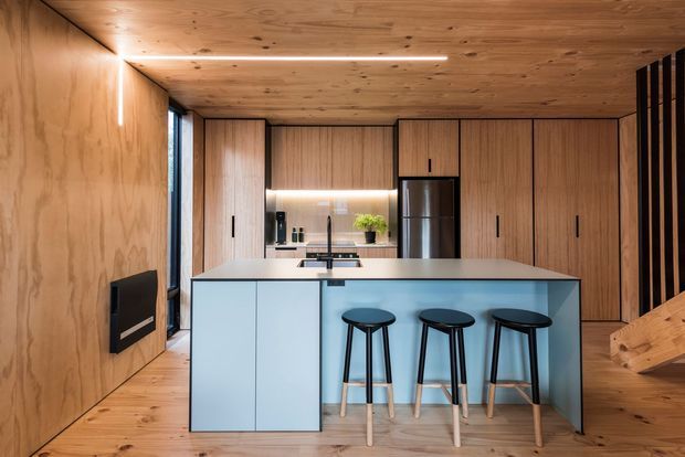 Compact modern kitchen clad extensively in warm timber veneer, with full-height timber cabinetry, a pale blue island bench, three bar stools, integrated appliances, and linear concealed lighting. A timber-lined ceiling and walls create a cohesive cabin-like interior, while a black slatted stair or screen element is visible at the right.