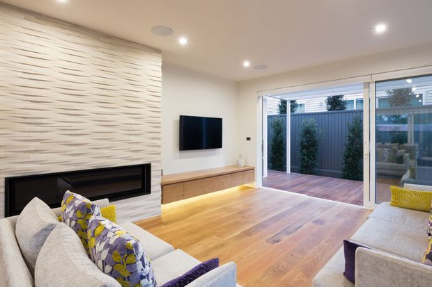 Contemporary living room with warm timber flooring, a large textured feature wall and built-in black fireplace, wall-mounted television, low timber media console, and substantial sliding glass doors opening to a private timber deck and landscaped courtyard. Neutral upholstered sofas with patterned cushions add colour and comfort.