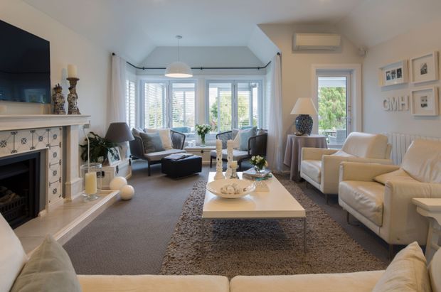Bright, traditionally styled living room with a large cream sectional sofa in the foreground, two upholstered armchairs, a central white coffee table, and a textured area rug. The room features a decorative fireplace with patterned tile surround and mantel, wall-mounted television, framed artwork, table lamps, pendant lighting, and extensive white-framed bay-style glazing with plantation shutters. A split-system air conditioner is mounted above a side window.