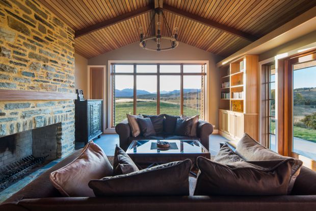 Rustic living room with a large rough-stone fireplace, exposed timber-lined vaulted ceiling, dark leather sofas and chairs, built-in timber shelving, and expansive windows overlooking a rural landscape. The warm materials and abundant natural light create a comfortable lodge-like setting.