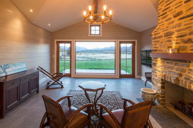 A warm, rustic living room with a pitched, timber-lined ceiling and horizontal timber wall panelling. The room features a large stone fireplace with a timber mantel, expansive divided glazing overlooking a rural landscape, a dark timber sideboard, leather lounge chairs, a patterned area rug, and a central chandelier.