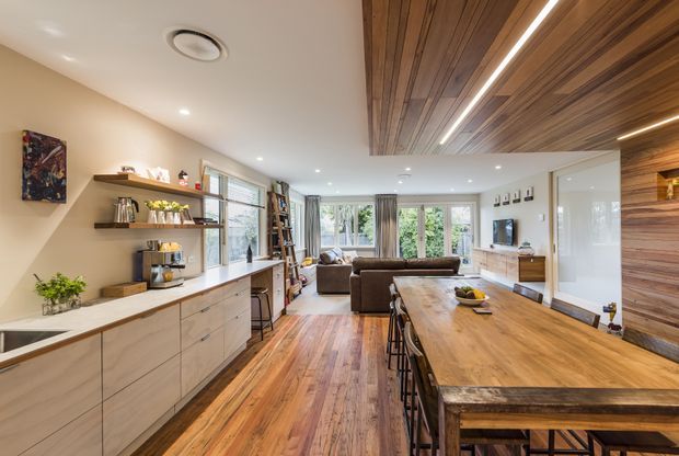 Open-plan residential interior combining a contemporary kitchen, dining and living area. The room features extensive warm timber flooring, a large timber dining table, pale built-in kitchen cabinetry with open shelving, a timber-lined ceiling feature, recessed lighting, and glazing opening toward greenery outside.
