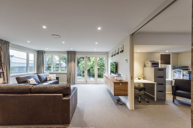 Bright open-plan living room with a large brown leather sectional, pale carpet, full-height windows and glazed doors opening to a garden. A timber wall-mounted media console and television sit along the right wall, with a compact home office visible through a wide opening beside it. The room has recessed ceiling lights, neutral finishes and soft curtains.