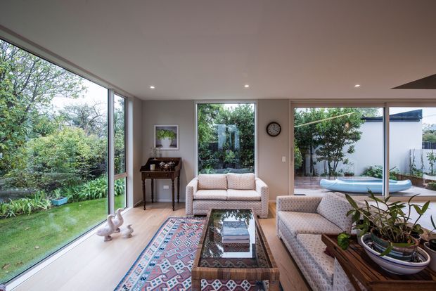 Bright contemporary living room with extensive floor-to-ceiling glazing overlooking a leafy garden and outdoor pool area. The room features pale timber flooring, light neutral walls, upholstered sofas and armchairs, a patterned area rug, a glass-topped coffee table, indoor plants, and recessed ceiling lighting.