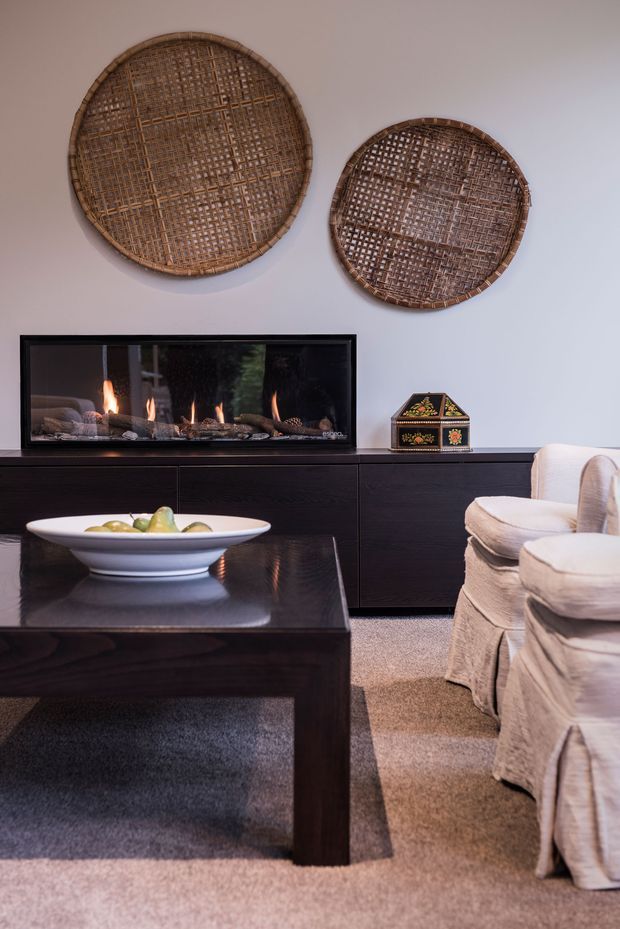 A refined living room with a dark timber media cabinet and matching square coffee table, a linear gas fireplace, and two large woven circular wall baskets above the fireplace. Upholstered cream armchairs sit beside a neutral carpet, while a white decorative bowl and patterned box add small accents. The room has soft warm lighting and a calm, contemporary-traditional feel.