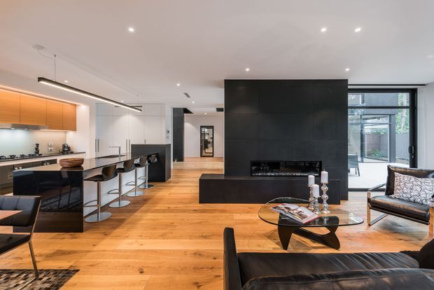 Open-plan contemporary interior combining a kitchen, dining area and living room. The space features warm timber flooring, a sleek black kitchen island and cabinetry, orange-toned wall cabinetry, a large dark feature wall with built-in fireplace, extensive glazing, modern recessed lighting and sculptural furniture. The room has a refined, high-contrast palette with a bright connection to an outdoor courtyard visible through the windows.